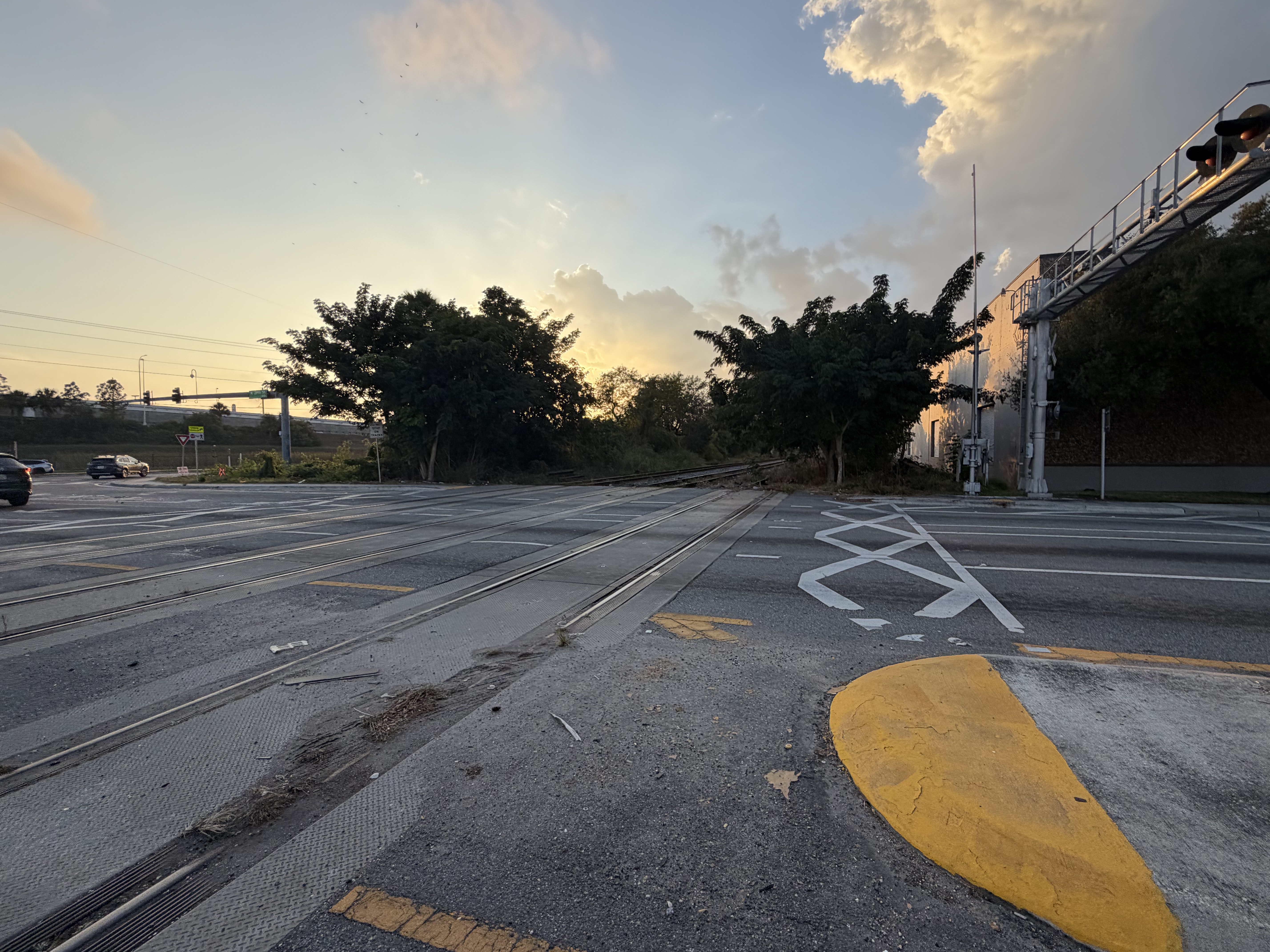 Road-level view of rail crossing with yellow lane markings, crossbuck marking, and elevated transit structure